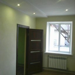 HUTMAN - Wnętrze pokoju po odświeżeniu: pomalowane ściany w odcieniach beżu, nowa podłoga imitująca drewno, białe okno z widokiem na metalową konstrukcję na zewnątrz, ciemne drzwi z trzema poziomymi...