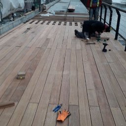 HUTMAN - Montaż drewnianego tarasu nad rzeką: pracownik na kolanach przykręca deski w pobliżu balustrady, drugi obserwuje, na podłodze leżą narzędzia i rękawice robocze.