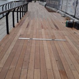 HUTMAN - Montaż desek tarasowych z drewna egzotycznego na nabrzeżu w Gdańsku, widoczna poziomica i narzędzia, w tle budynki i rzeka.