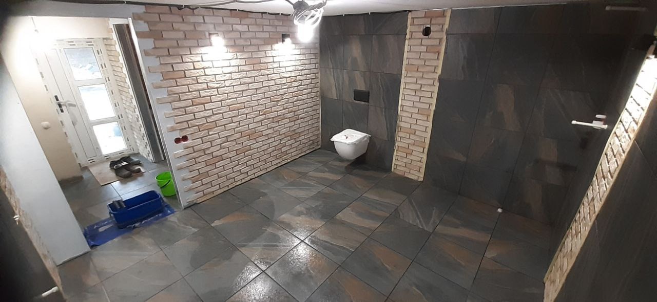 Wnętrze łazienki w trakcie remontu, widoczne płytki imitujące cegłę na ścianie, ciemne płytki na podłodze i ścianach, częściowo zamontowana toaleta, oraz akcesoria malarskie i wiadro w rogu...