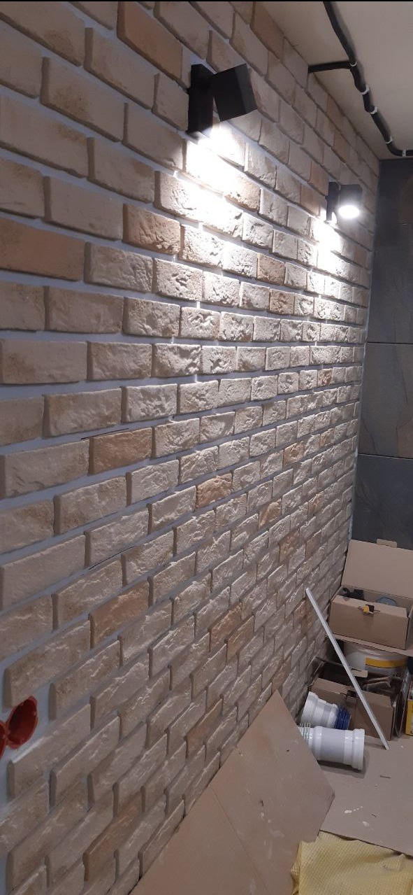 Ściana wykończona cegiełkami klinkierowymi w odcieniach beżu i brązu, oświetlona dwoma czarnymi kinkietami, z widocznymi elementami instalacji elektrycznej i materiałami budowlanymi w tle.