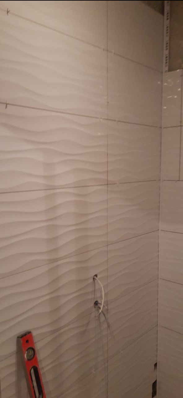 Układanie białych, falistych płytek w łazience, widoczny poziomica i wystające kable z otworu w ścianie.