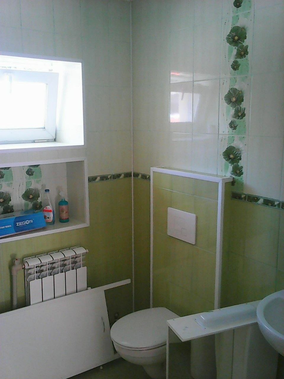 Zielono-biała łazienka z płytkami w kwiaty, widoczna toaleta, umywalka, grzejnik i półka z kosmetykami.