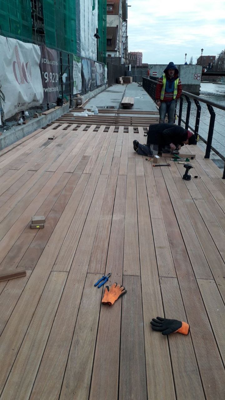 Montaż drewnianego tarasu nad rzeką: pracownik na kolanach przykręca deski w pobliżu balustrady, drugi obserwuje, na podłodze leżą narzędzia i rękawice robocze.