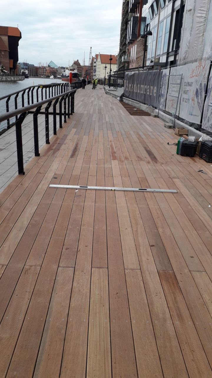 Montaż desek tarasowych z drewna egzotycznego na nabrzeżu w Gdańsku, widoczna poziomica i narzędzia, w tle budynki i rzeka.