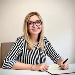 Uśmiechnięta blondynka w okularach i bluzce w czarno-białe paski, siedząca za biurkiem i pisząca czarnym piórem w otwartym notatniku z czerwonymi brzegami.