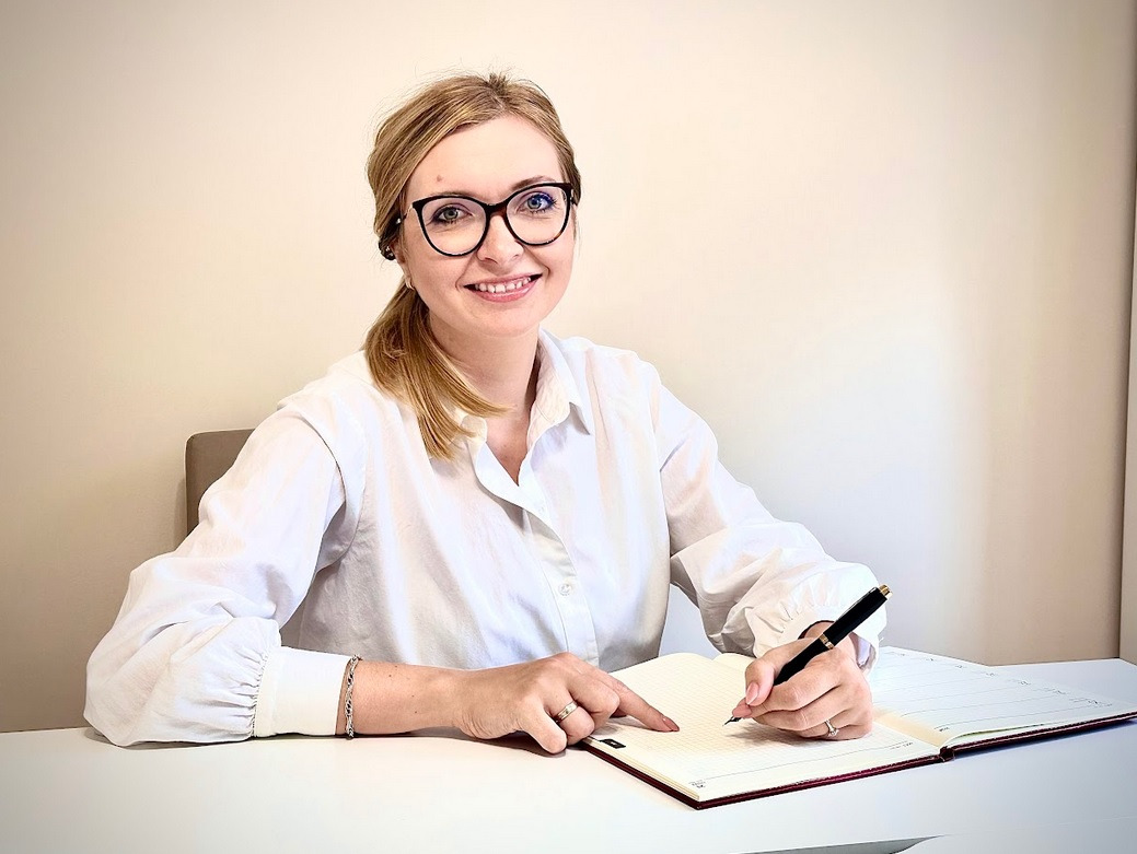 Uśmiechnięta blondynka w okularach siedzi przy biurku, pisząc w otwartym kalendarzu piórem wiecznym.