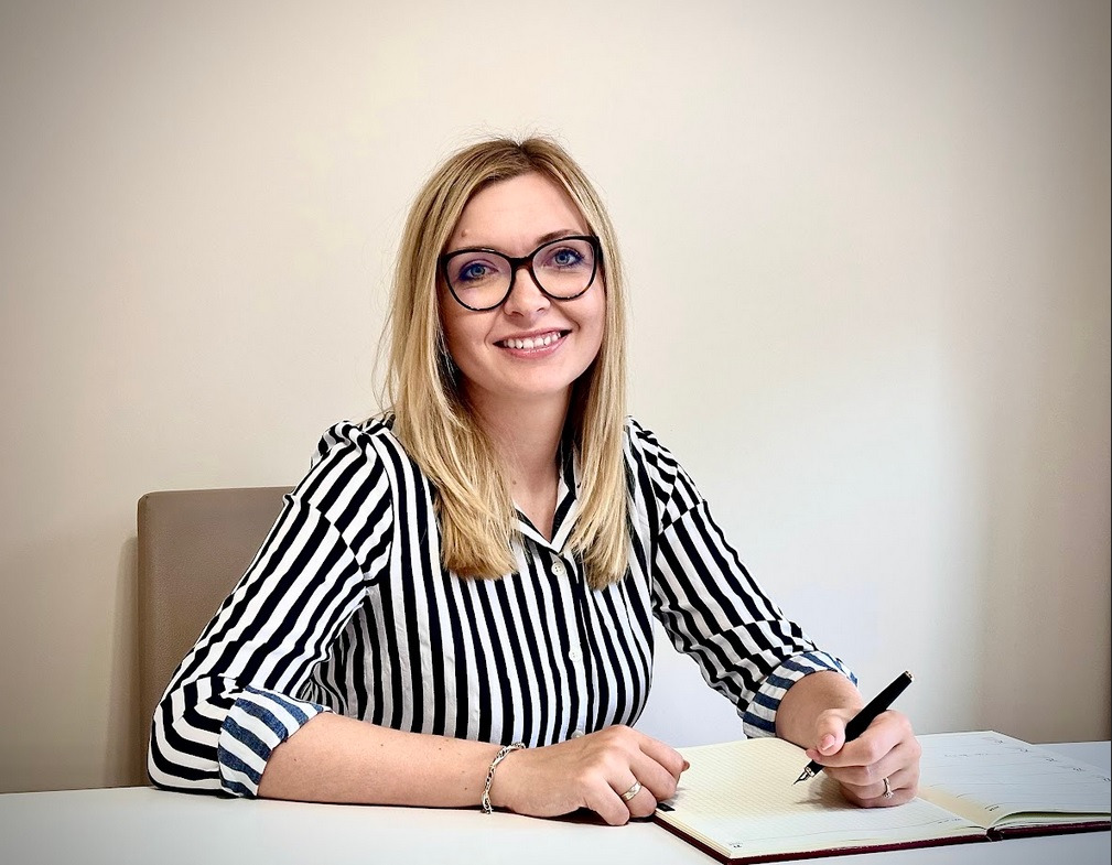 Uśmiechnięta blondynka w okularach i bluzce w czarno-białe paski, siedząca za biurkiem i pisząca czarnym piórem w otwartym notatniku z czerwonymi brzegami.