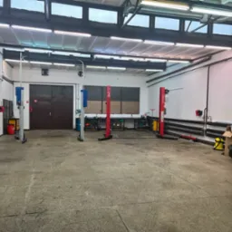 Przestronny warsztat samochodowy z dwoma podnośnikami kolumnowymi w kolorach czerwonym i niebieskim, widoczne narzędzia i wyposażenie warsztatowe, posadzka betonowa. Widok na bramę wjazdową.