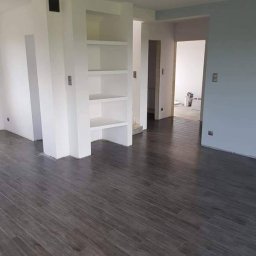 Piotr Góralczuk - Nowoczesne wnętrze po remoncie z ciemną, drewnopodobną podłogą, białe ściany z wnęką na regał i widoczne przejścia do innych pomieszczeń. W tle wiadro z farbą.