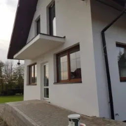 Świeżo pomalowana elewacja domu jednorodzinnego z brązowymi oknami, widoczne trzy wiadra farby u podstawy ściany.