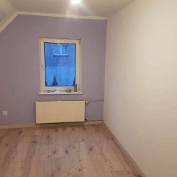 Piotr Góralczuk - Pomalowane na liliowo pomieszczenie z drewnianą podłogą i białym grzejnikiem pod oknem, jedna ściana biała