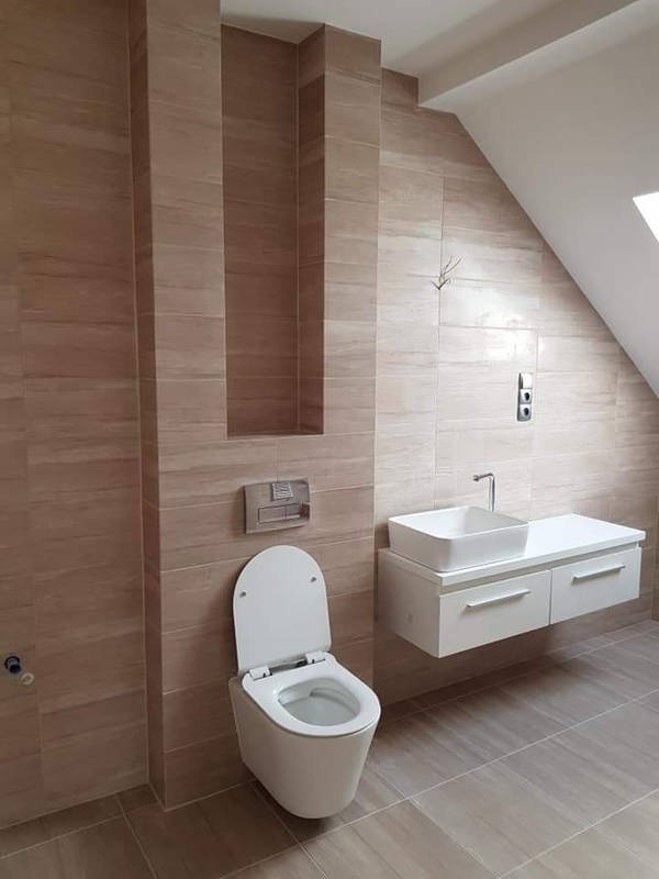 Nowoczesna łazienka w beżowych płytkach imitujących drewno, z podwieszaną toaletą i umywalką na szafce, w pomieszczeniu ze skosem.