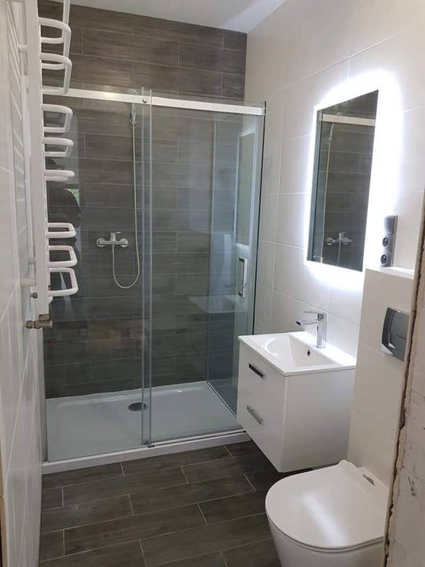 Nowoczesna łazienka z kabiną prysznicową z przesuwnymi drzwiami, podświetlanym lustrem, umywalką z szafką i toaletą, wykończona płytkami imitującymi drewno i białymi płytkami.