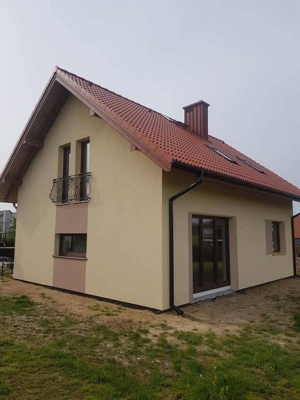 Otynkowany dom jednorodzinny z brązową dachówką, widok z boku na świeżo położoną elewację w odcieniach beżu i brązu.