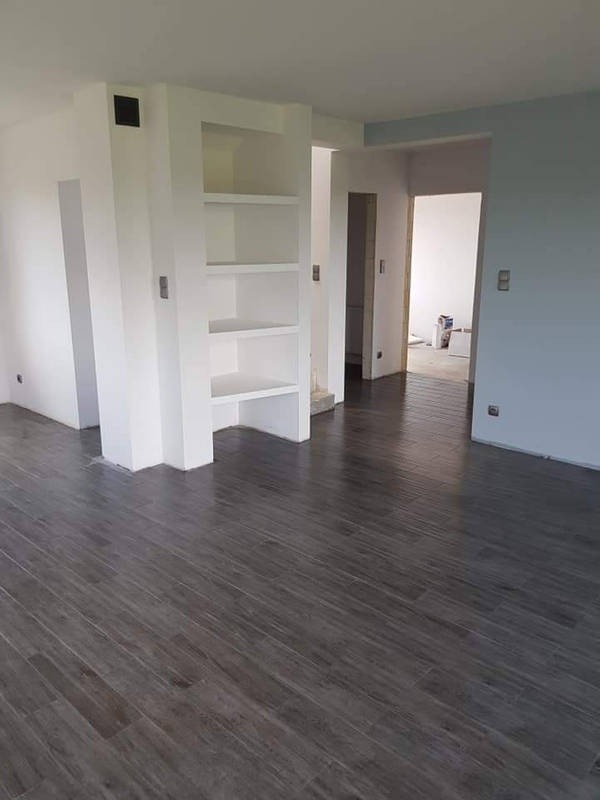 Nowoczesne wnętrze po remoncie z ciemną, drewnopodobną podłogą, białe ściany z wnęką na regał i widoczne przejścia do innych pomieszczeń. W tle wiadro z farbą.