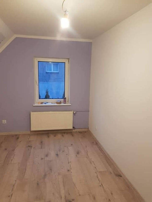 Pomalowane na liliowo pomieszczenie z drewnianą podłogą i białym grzejnikiem pod oknem, jedna ściana biała