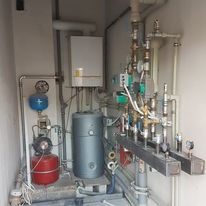 Kompaktowa kotłownia z piecem, zasobnikiem ciepłej wody i rozbudowaną instalacją hydrauliczną w małym pomieszczeniu technicznym. Widoczne liczne zawory, pompy obiegowe i manometry.