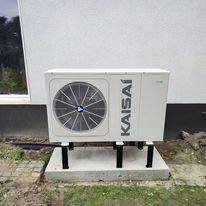 Biała pompa ciepła Kaisai zamontowana na betonowym fundamencie, z widoczną zewnętrzną jednostką wentylacyjną i fragmentem białej ściany budynku w tle.