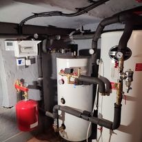 Zestaw urządzeń grzewczych w piwnicy: biały zasobnik ciepłej wody z manometrami, czerwony zbiornik wyrównawczy, szare rury izolowane i panel sterowania na ścianie.