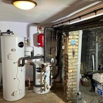 Białe zbiorniki akumulacyjne ciepła z czarnymi rurami w piwnicy z odsłoniętą cegłą i instalacją rurową na ścianie, oświetlone lampą sufitową.