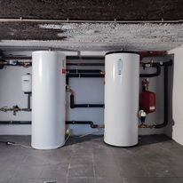 Dwa białe, cylindryczne zbiorniki na wodę ustawione obok siebie w pomieszczeniu technicznym z betonowym sufitem i licznymi rurami. Widoczny czerwony zbiornik wyrównawczy i inne elementy instalacji.