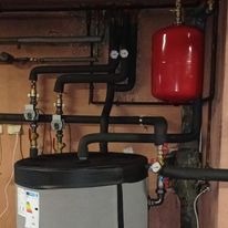 Szary zbiornik buforowy z czarną pokrywą, połączony rurami w izolacji termicznej, czerwony zbiornik wyrównawczy, manometry i armatura w kotłowni o ciemnych ścianach.