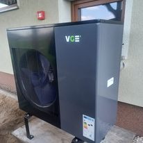 Zewnętrzna jednostka pompy ciepła VGE w kolorze grafitowym, zamontowana na metalowych wspornikach przy jasnej ścianie budynku. Widoczny wentylator i etykieta energetyczna.