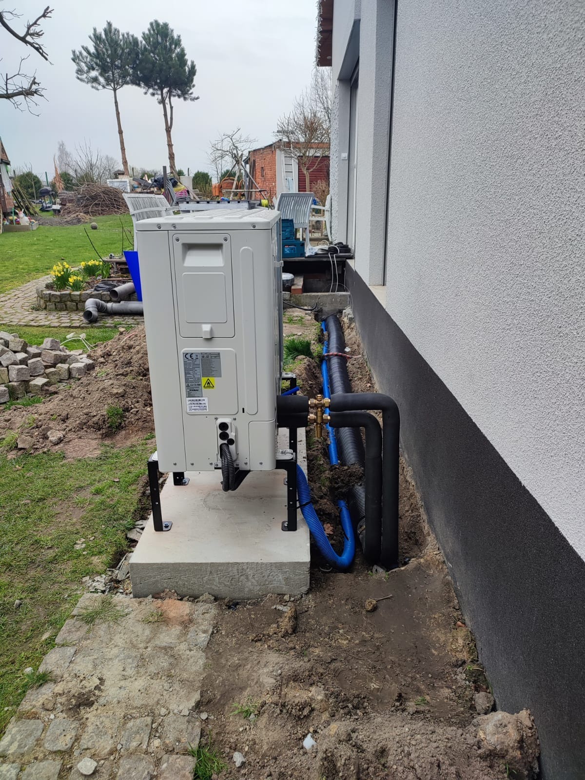 Biała jednostka zewnętrzna pompy ciepła, zamontowana na betonowej podstawie obok jasnoszarej ściany budynku, z widocznymi czarnymi i niebieskimi rurami instalacyjnymi w ziemi.
