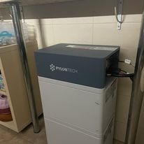 Akumulator Pylontech umieszczony na białej szafce w rogu pomieszczenia, widoczne kable podłączone do urządzenia, fragment stołu u góry kadru.