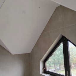 Bartosz Stec - Wykończone wnętrze w Domaradzu z płytami kartonowo-gipsowymi na skośnym suficie i ścianach wyłożonych beżowymi płytkami, trójkątne okno z widokiem na ogród.