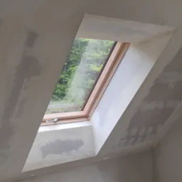 Poddasze ze skośnym oknem dachowym w trakcie szpachlowania ścian. Widoczne ślady po łatach gipsowych na szarej powierzchni, okno z widokiem na zieleń.