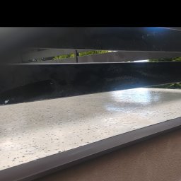 Bartosz Stec - Jasna, nakrapiana powierzchnia tarasu lub balkonu, z widocznym odbiciem światła, otoczona czarną, metalową balustradą z poziomymi elementami, na tle zielonej roślinności.