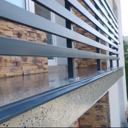 Bartosz Stec - Szara, metalowa balustrada balkonu z poziomymi profilami, obok ściany wykończonej beżowym kamieniem elewacyjnym i błyszczącą, nakrapianą posadzką.