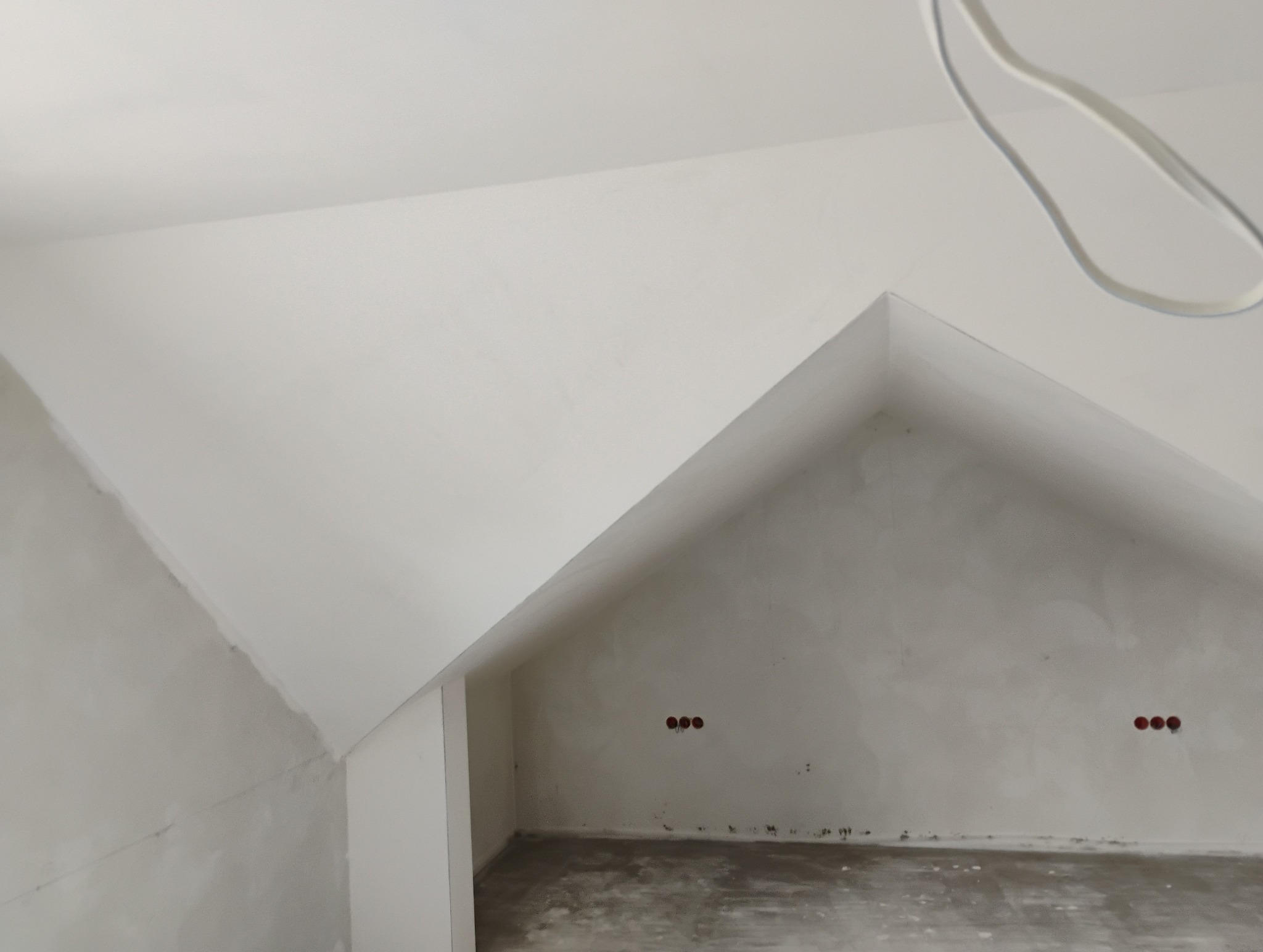 Poddasze w trakcie prac wykończeniowych, widoczne surowe ściany i fragmenty pomalowanej powierzchni, instalacja elektryczna wystaje ze ściany.