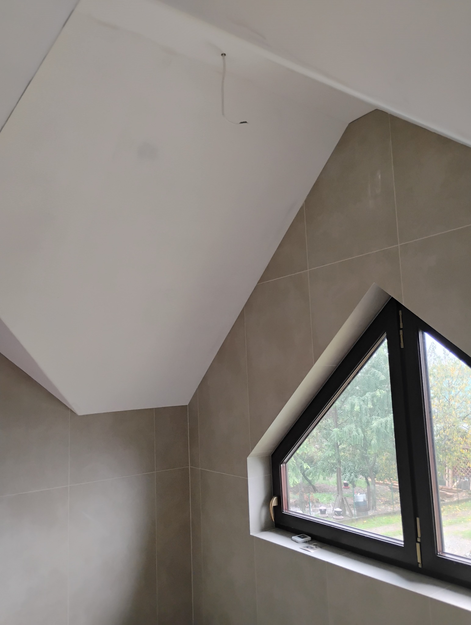 Wykończone wnętrze w Domaradzu z płytami kartonowo-gipsowymi na skośnym suficie i ścianach wyłożonych beżowymi płytkami, trójkątne okno z widokiem na ogród.