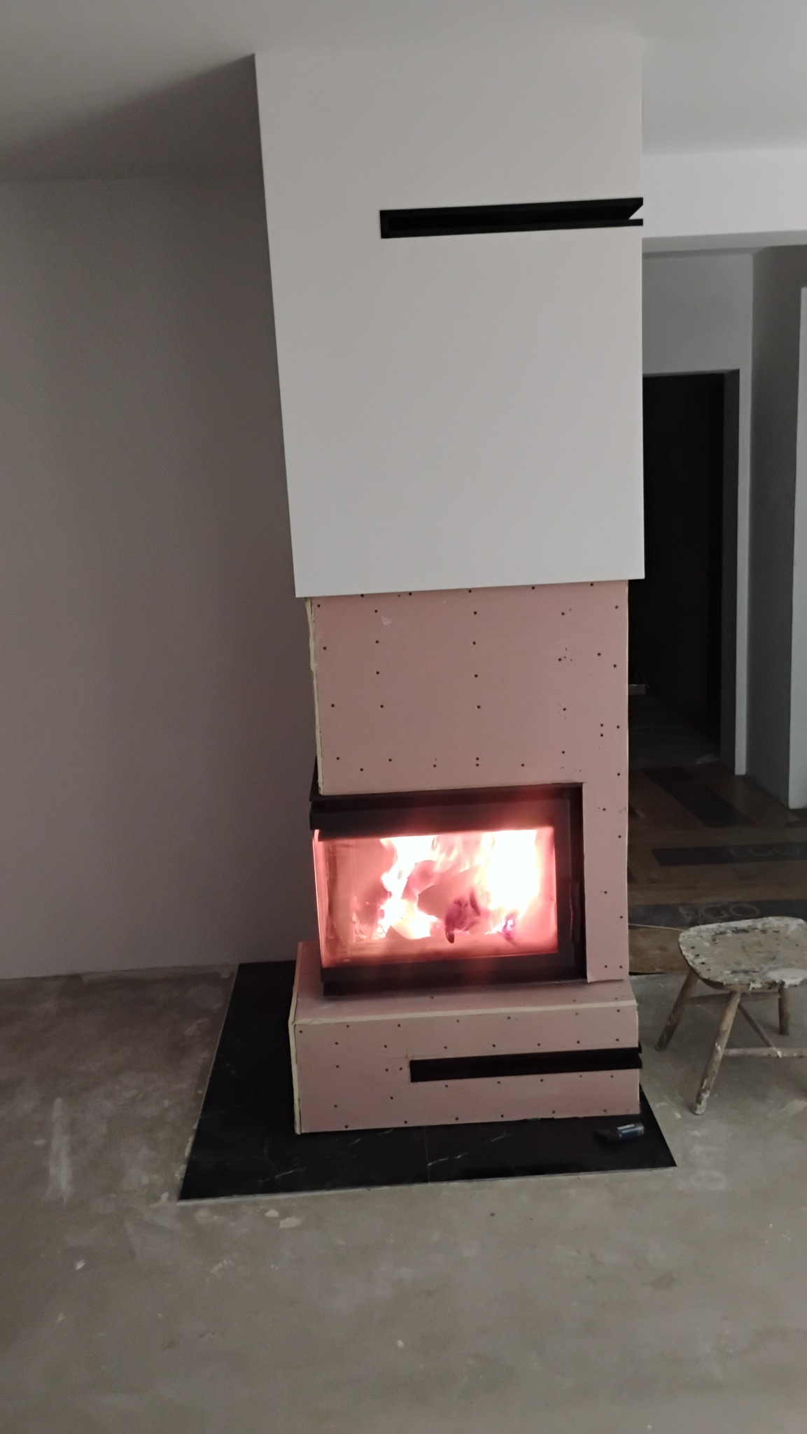 Wnętrze z kominkiem w trakcie obudowy z płyty kartonowo-gipsowej, widoczne płomienie i fragment krzesła.