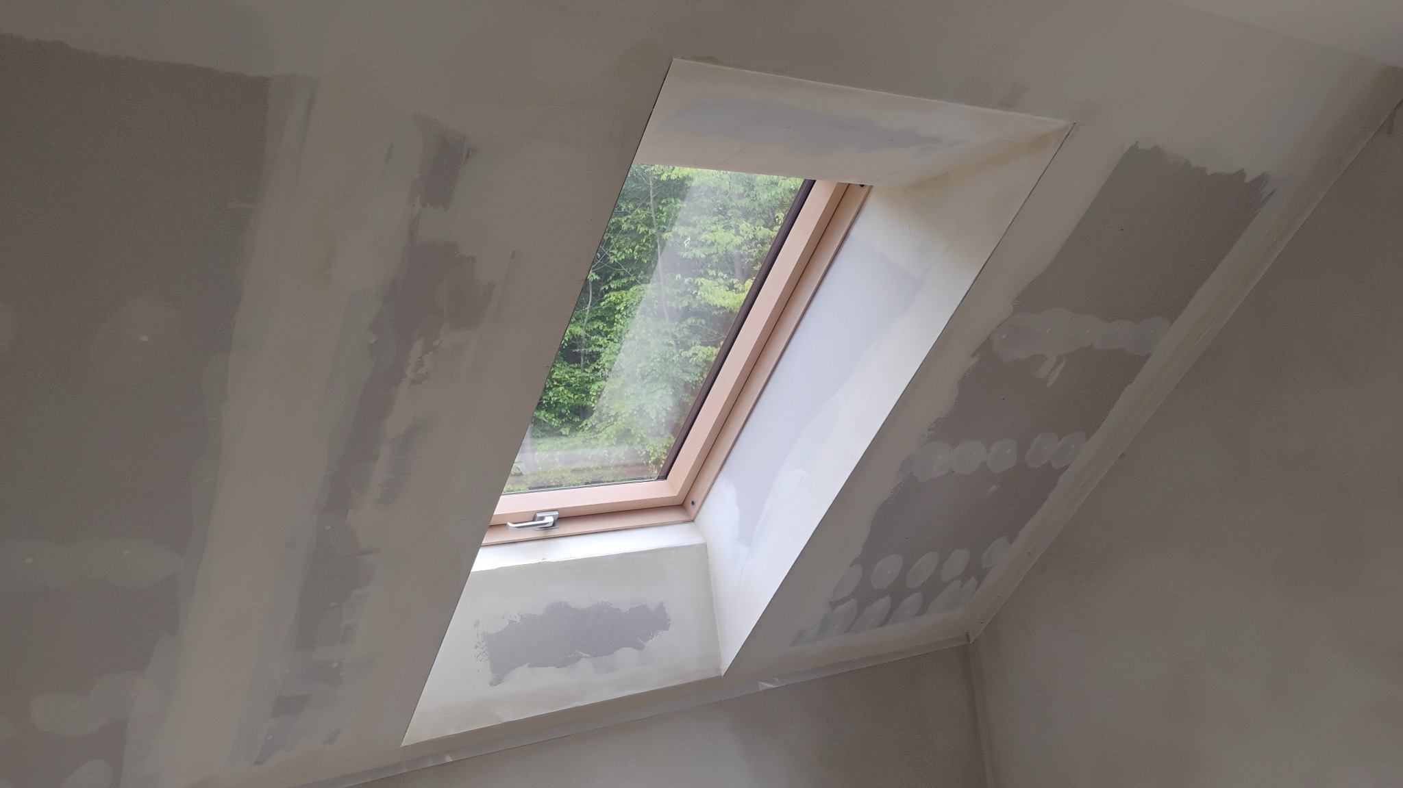 Poddasze ze skośnym oknem dachowym w trakcie szpachlowania ścian. Widoczne ślady po łatach gipsowych na szarej powierzchni, okno z widokiem na zieleń.