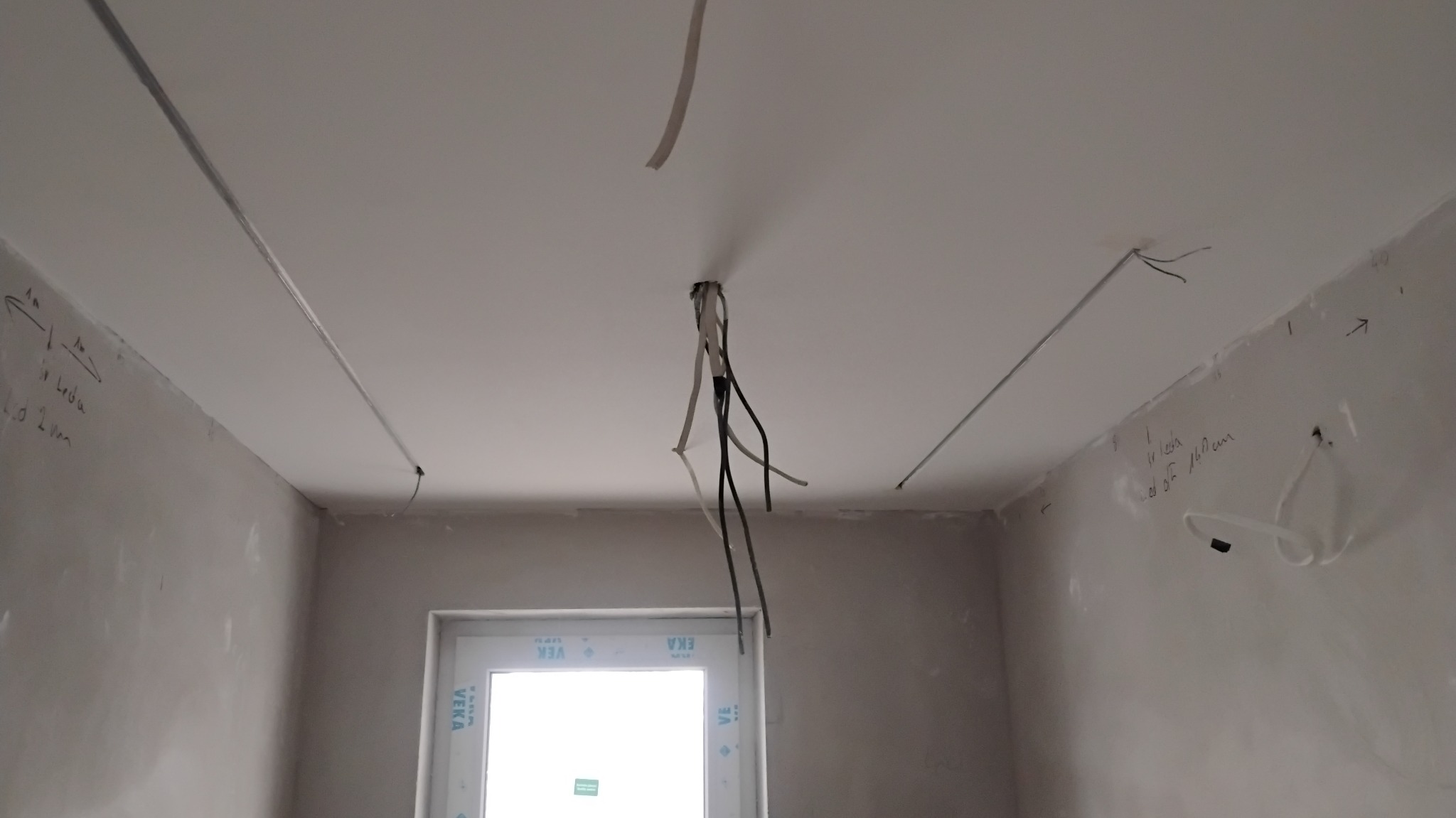 Surowy stan wnętrza: widoczne kable zwisające z sufitu, profile LED, ściany przygotowane do gładzi z odręcznymi notatkami, okno z PCV. Etap prac wykończeniowych.