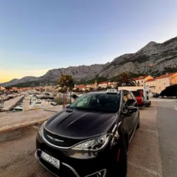 Czarny samochód taxi na tle malowniczego krajobrazu z górami i portem jachtowym. Widoczna tablica rejestracyjna SCI 7518L.