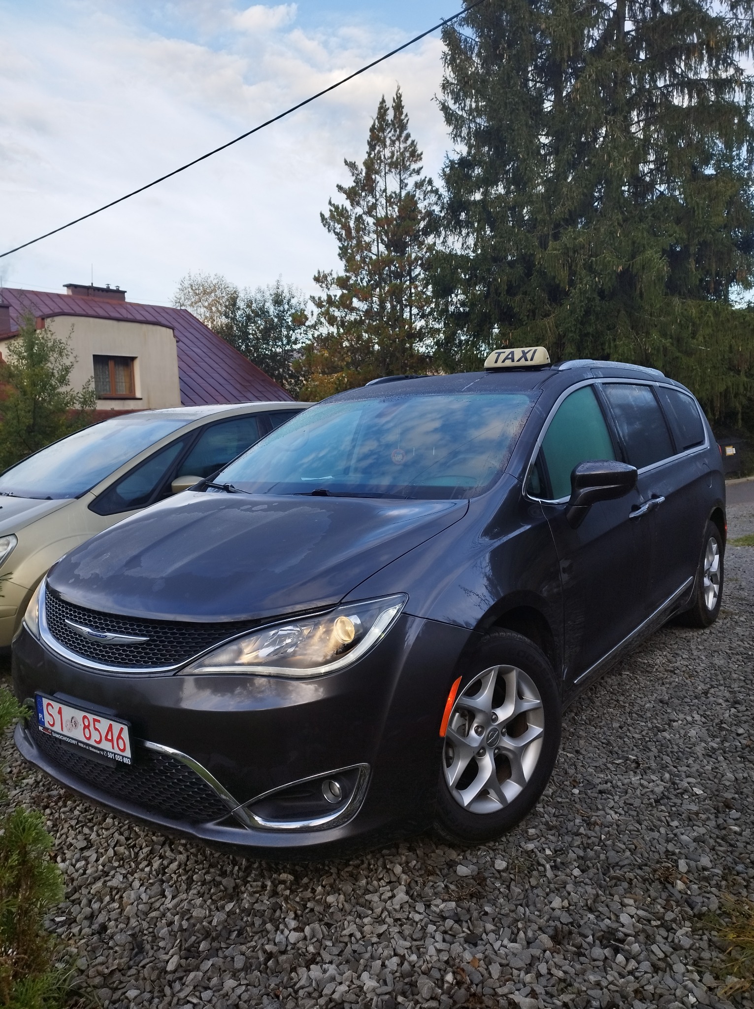 Ciemny minivan z napisem 'TAXI' na dachu, zaparkowany na żwirowym podjeździe, obok stoi jasny samochód osobowy, w tle dom i drzewa.
