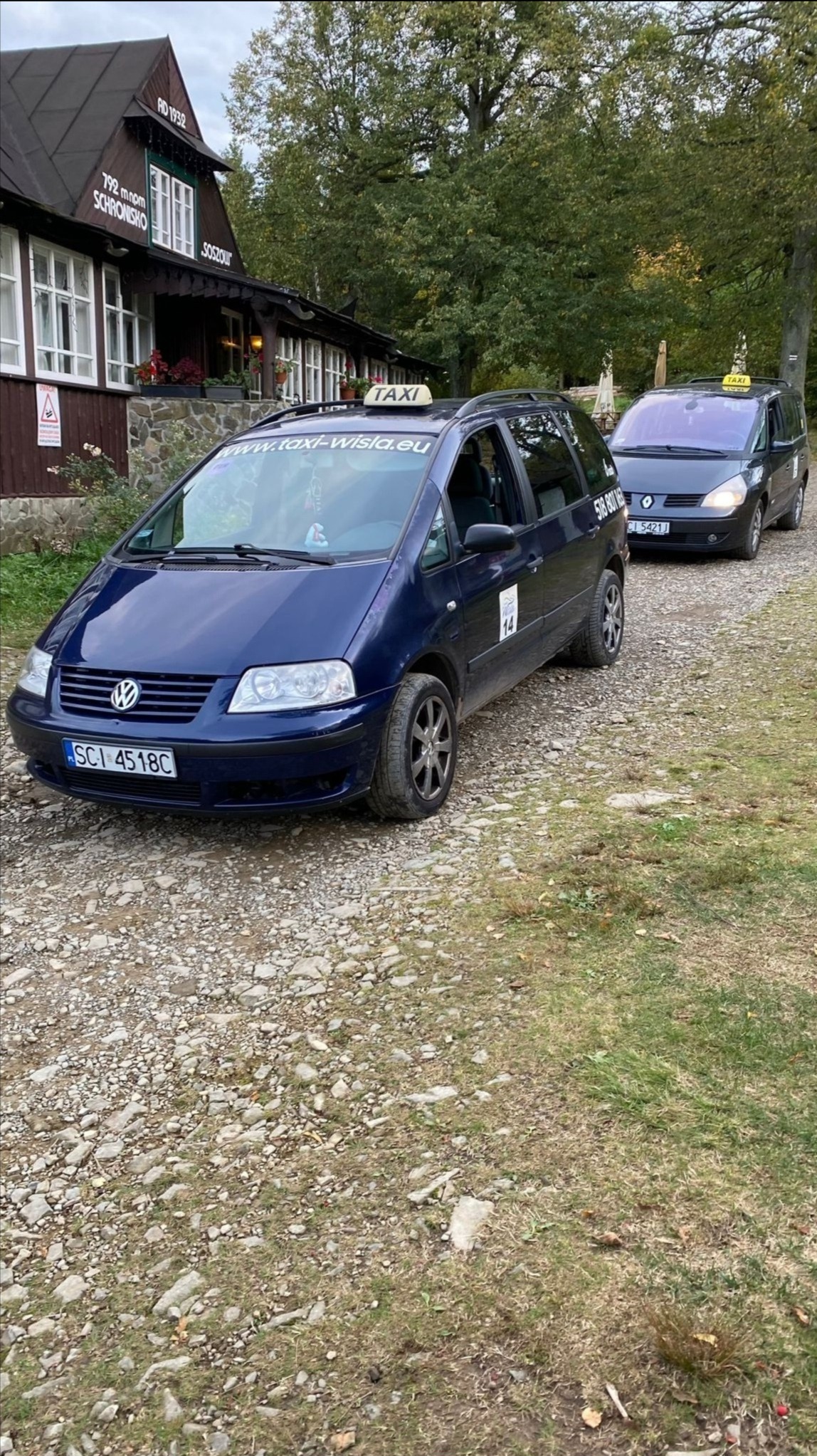 Dwa samochody taxi (VW Sharan i Renault Espace) zaparkowane na żwirowej drodze przed górskim schroniskiem Soszów w Wiśle. Na dachu każdego auta widoczny napis TAXI.