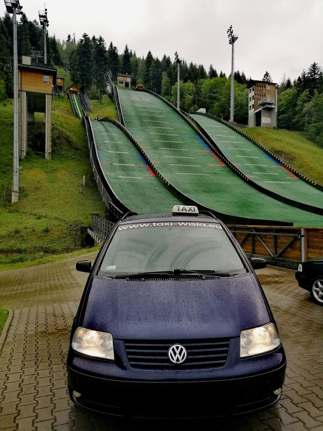 Niebieski samochód taxi marki Volkswagen Sharan z napisem www.taxi-wisla.eu na przedniej szybie, zaparkowany na tle skoczni narciarskiej w Wiśle.