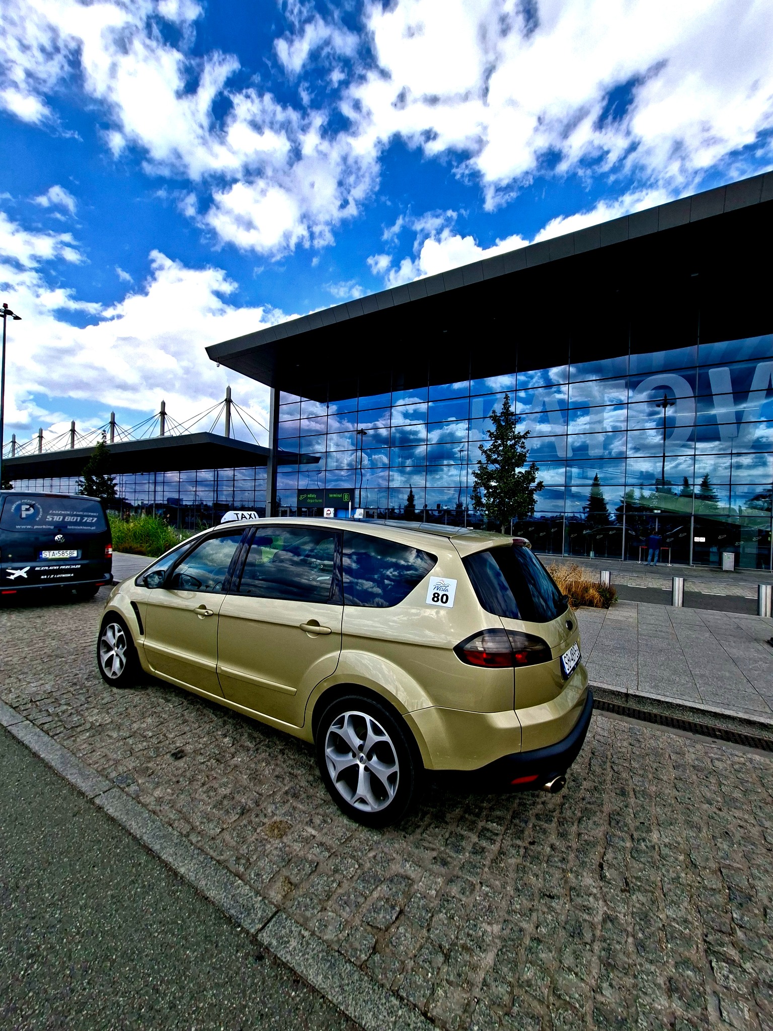 Złoty samochód typu kombi z napisem TAXI na dachu, zaparkowany przed nowoczesnym budynkiem lotniska z szklaną fasadą odbijającą błękitne niebo z chmurami.