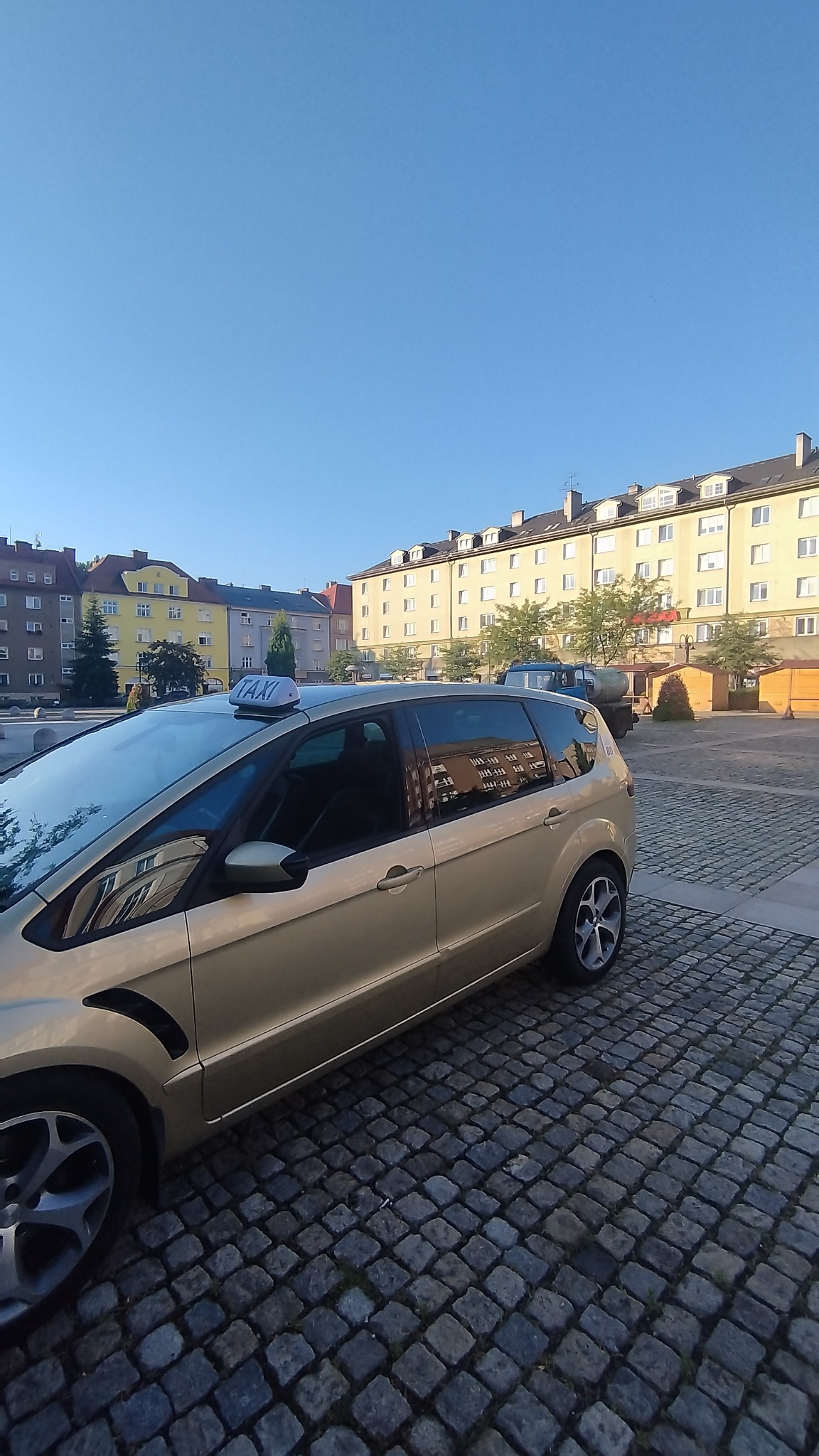 Złoty samochód typu van z napisem TAXI na dachu, zaparkowany na brukowanym placu miejskim w słoneczny dzień. W tle widoczne budynki mieszkalne i drzewa.