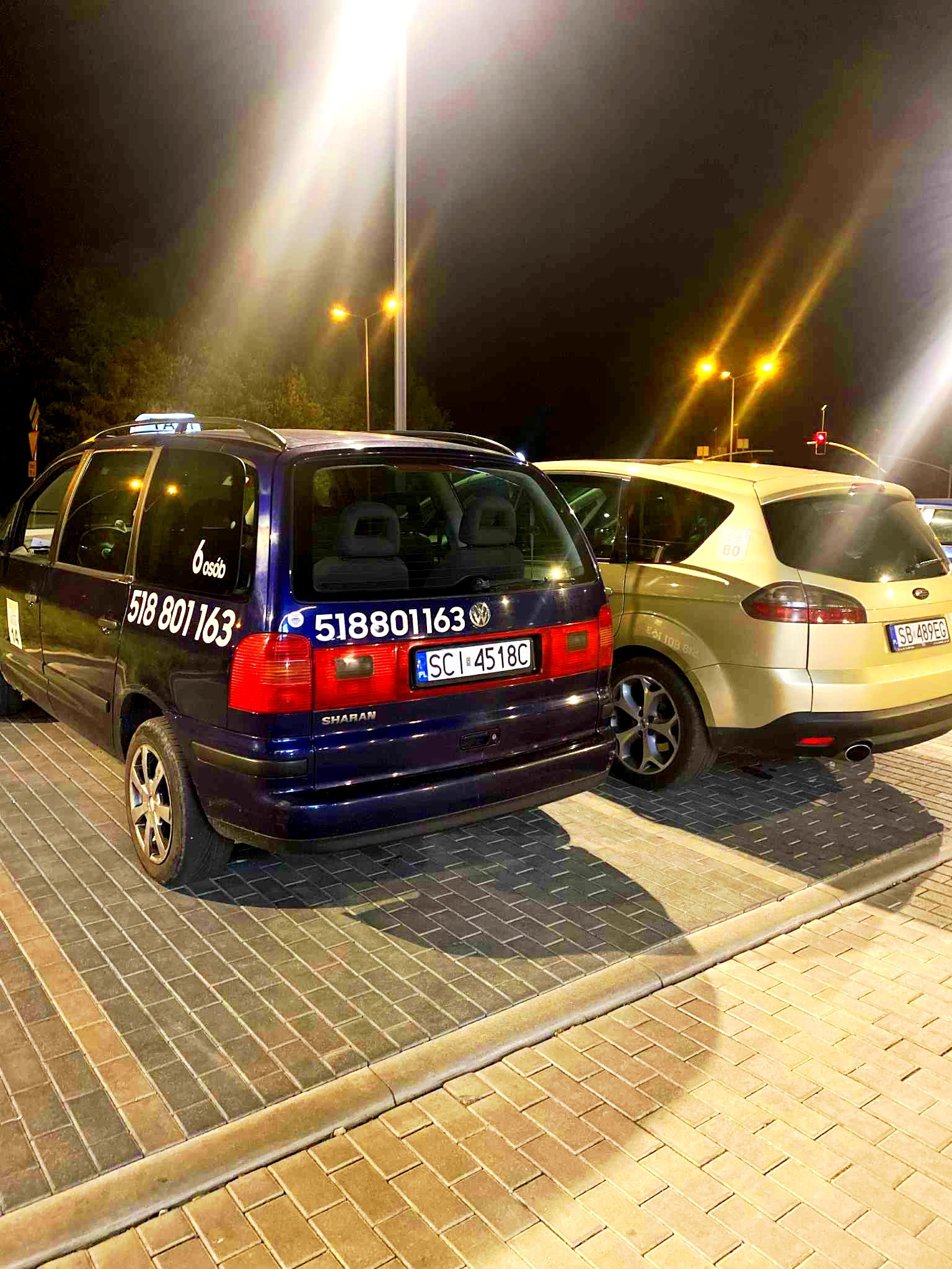 Dwa busy na parkingu nocą: granatowy VW Sharan z oznaczeniem taxi (6 osób) i złotawy Ford S-Max z numerem 80. Widoczne numery telefonów i tablice rejestracyjne.