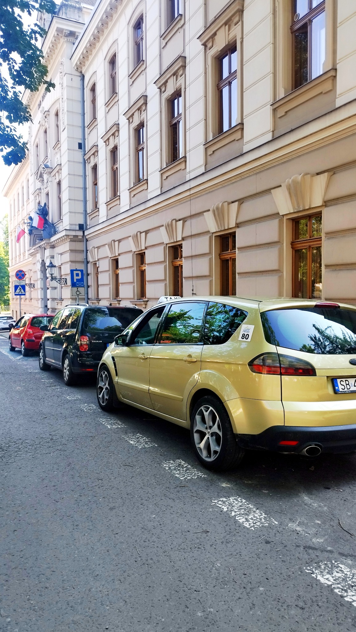 Żółty samochód osobowy Ford S-Max z napisem TAXI na dachu, zaparkowany na ulicy w mieście, na tle budynku z licznymi oknami.