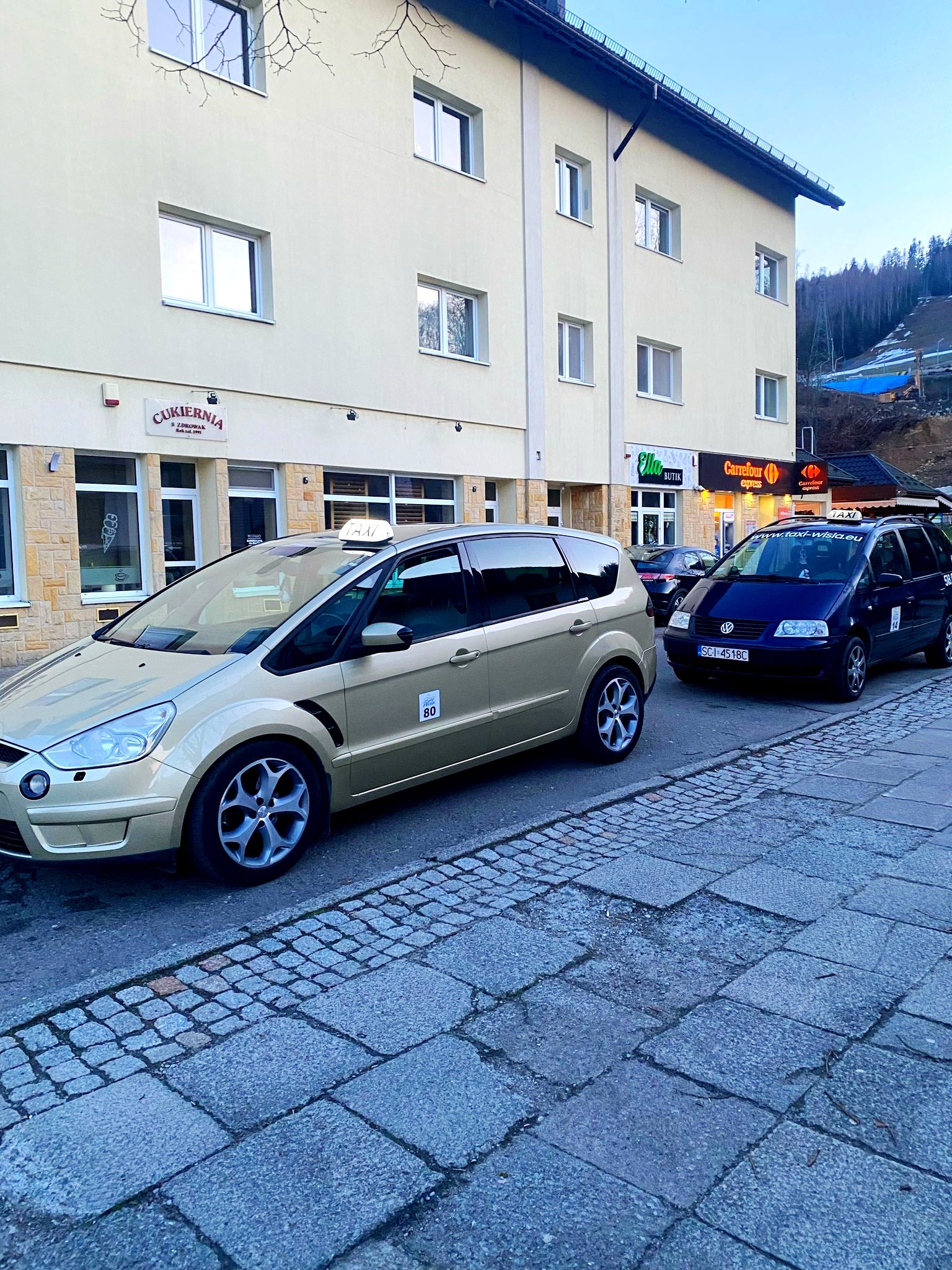 Dwa samochody taxi, złoty Ford S-Max z numerem bocznym 80 i granatowy Volkswagen Sharan, zaparkowane na ulicy w Wiśle, z widocznymi szyldami sklepów w tle.