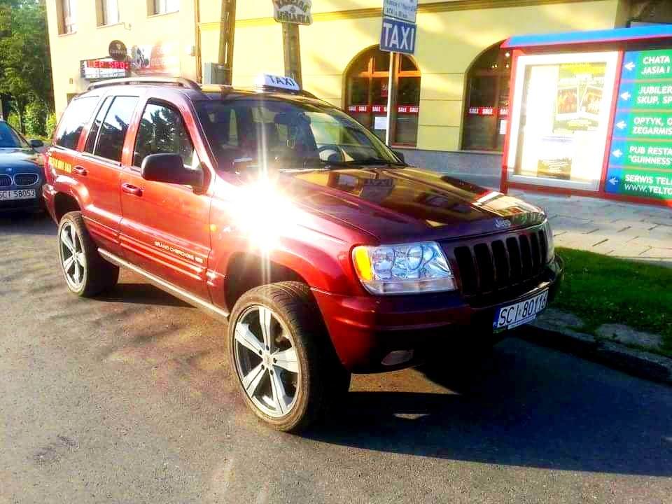 Czerwony Jeep Grand Cherokee z oznaczeniem Taxi na dachu, zaparkowany na ulicy w słoneczny dzień. Widoczny fragment budynku i tablica informacyjna.