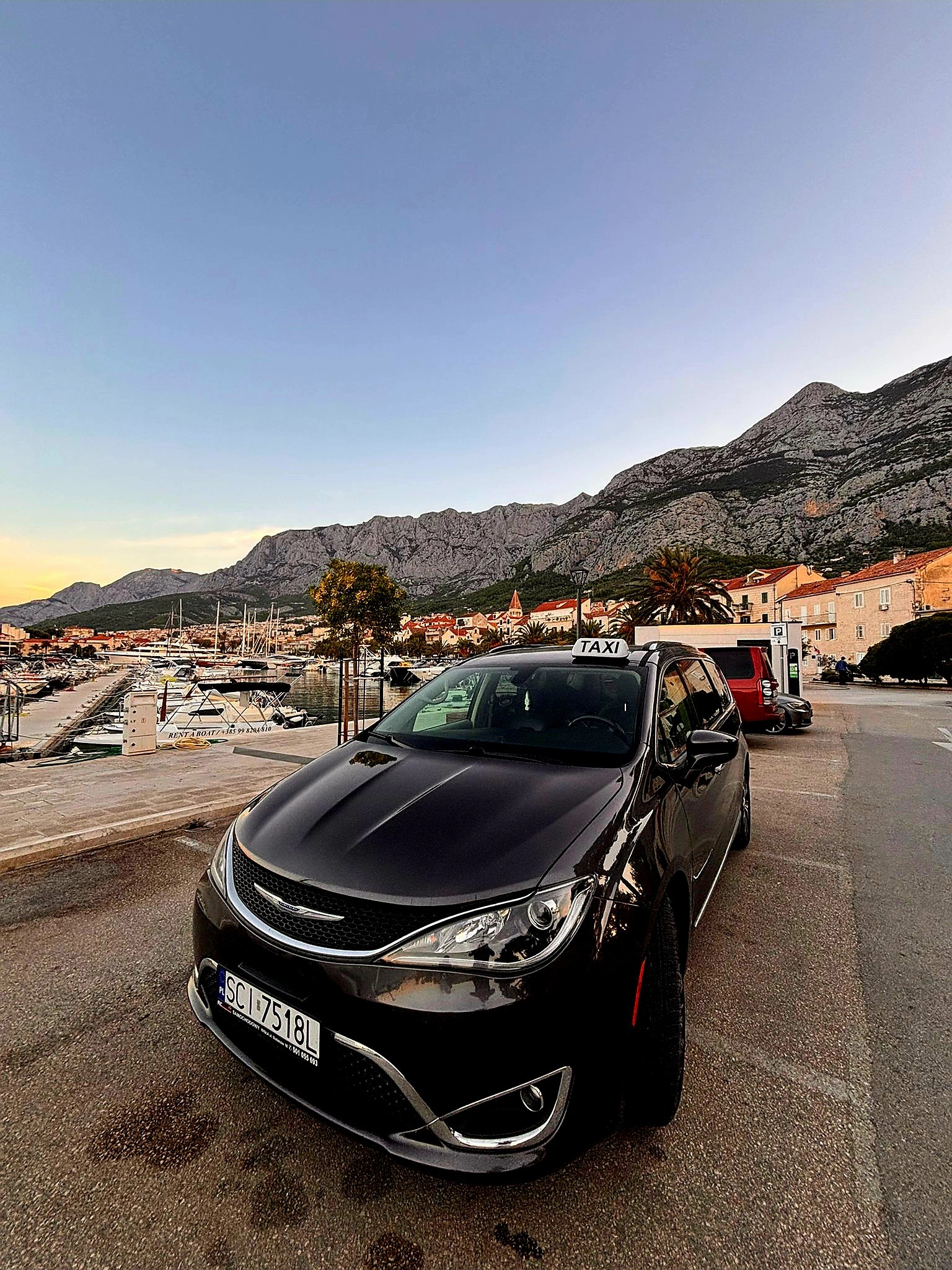 Czarny samochód taxi na tle malowniczego krajobrazu z górami i portem jachtowym. Widoczna tablica rejestracyjna SCI 7518L.